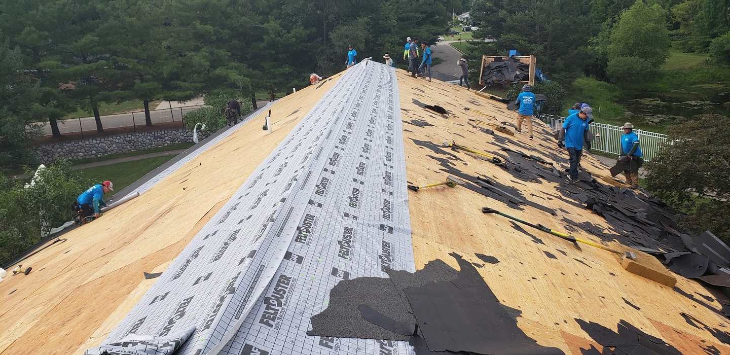 A crew in blue shirts stripping old shingles from a large roof, with a strip of new synthetic underlayment rolled up the ridge and pine trees behind.
