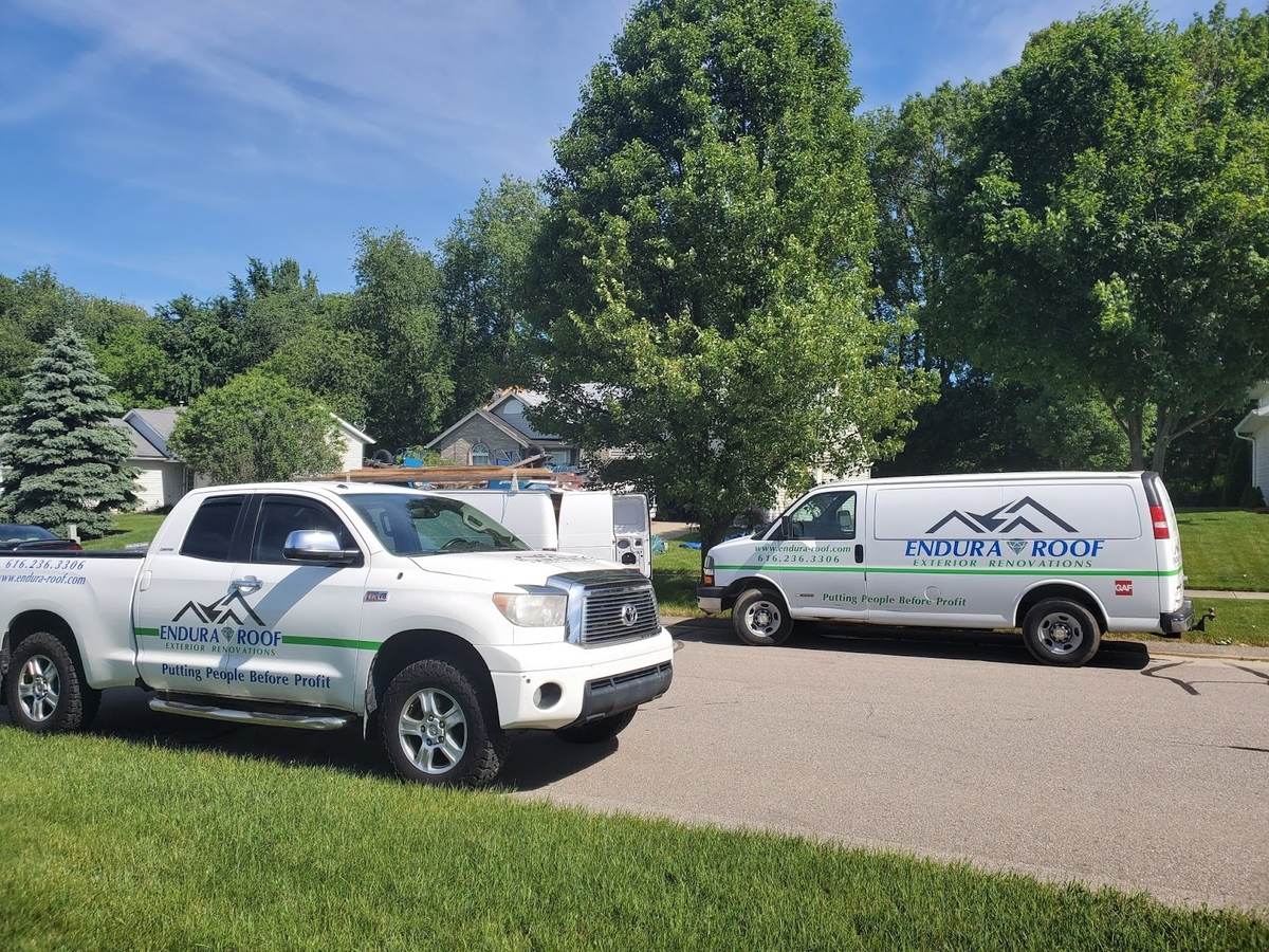 A white Endura-Roof pickup and a white Endura-Roof van, both lettered with a mountain-peak logo and a green stripe, parked on a residential street under tall trees.