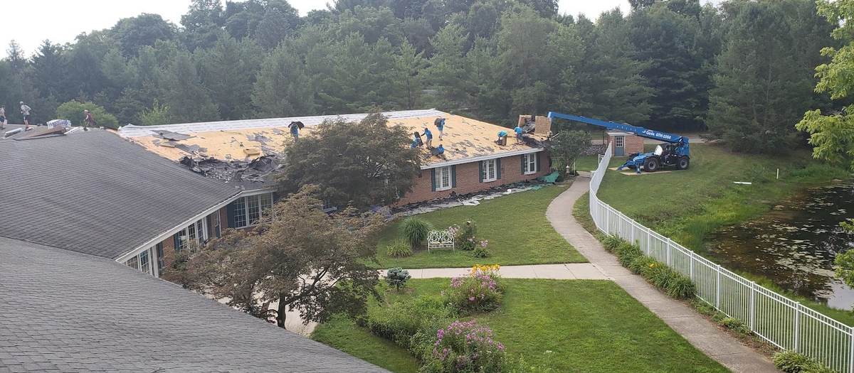 An aerial view of a long brick building with half the roof stripped to bare decking, a crew working on it, and a blue telehandler parked on the lawn beside a white fence.